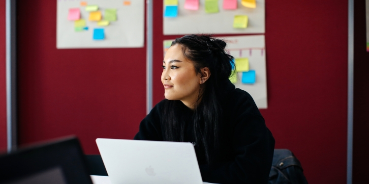 A photograph of a lady sitting at a desk, behind a laptop, gazing to the side and smiling. In the background there is a red wall with planners and post-its stuck on.